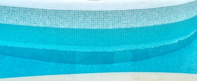 A close-up view of a curved swimming pool edge with a mosaic tile pattern. The tiles are primarily blue with varying shades, creating a gradient effect in the water. The upper portion of the pool is lined with white tiles, and the pool water appears clean and turquoise.