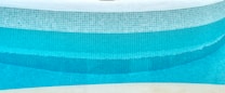 A close-up view of a curved swimming pool edge with a mosaic tile pattern. The tiles are primarily blue with varying shades, creating a gradient effect in the water. The upper portion of the pool is lined with white tiles, and the pool water appears clean and turquoise.