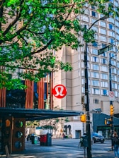 A bustling city street corner with a large, prominent red and white logo on a building, likely related to a retail store. In the foreground, lush green tree branches provide shade. The street signs read Burrard St and Robson St, indicating a well-known urban location. Various vehicles and pedestrians are visible, contributing to the lively atmosphere.