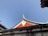 A traditional architectural roof with an ornate curved design and a bright red triangular pattern beneath. The structure is set against a clear blue sky, with some weathered textures visible on the surrounding walls.