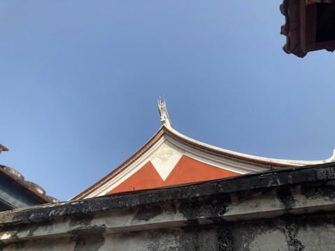 A traditional architectural roof with an ornate curved design and a bright red triangular pattern beneath. The structure is set against a clear blue sky, with some weathered textures visible on the surrounding walls.