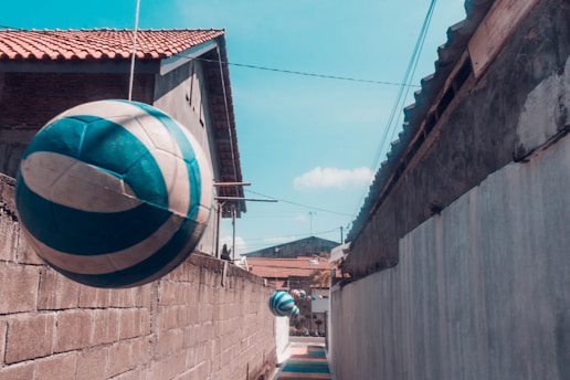A narrow alleyway lined with concrete and brick walls, featuring several blue and white volleyballs suspended on strings above the walkway. The walls are topped with rooftops, and the sky is clear with a few clouds.