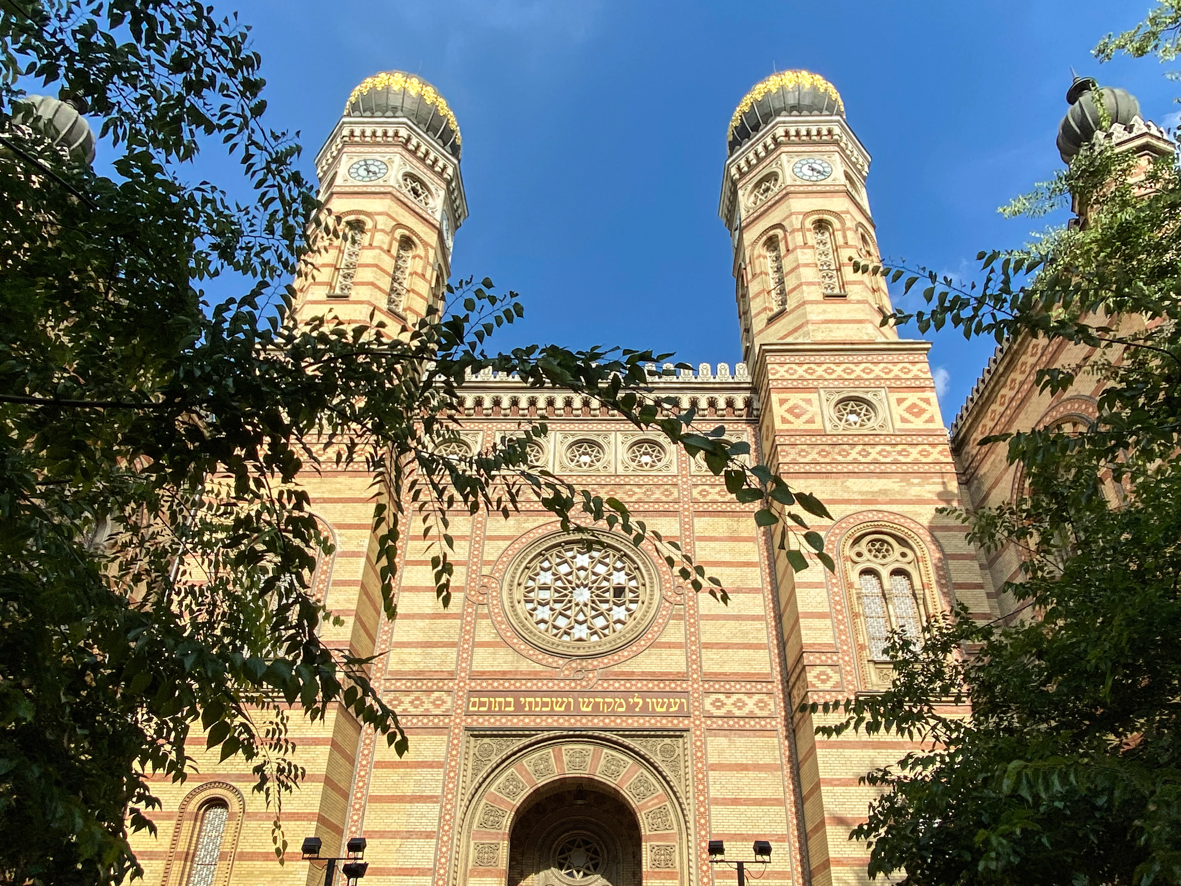 The twin onion-domed towers and stone rose window of the Dohany Street Synagogue in Budapest's Jewish Quarter, its striped brick and gilded pattern glowing against a deep blue sky behind summer leaves