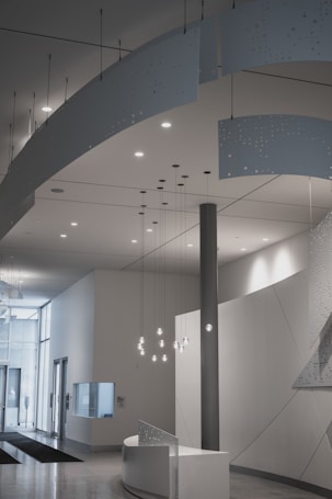 A modern interior space featuring a minimalist design with clean lines and neutral colors. Suspended decorative panels with perforated patterns hang from the ceiling, and a set of pendant lights illuminate the area. The room includes a curved reception desk, and large glass doors lead to an outside view, allowing natural light to fill the area.