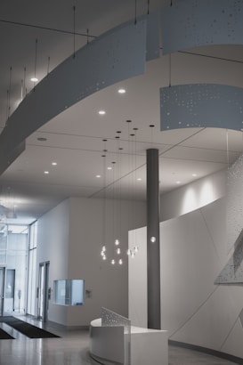 A modern interior space featuring a minimalist design with clean lines and neutral colors. Suspended decorative panels with perforated patterns hang from the ceiling, and a set of pendant lights illuminate the area. The room includes a curved reception desk, and large glass doors lead to an outside view, allowing natural light to fill the area.