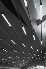 A series of modern, linear ceiling lights are suspended in an architectural interior. The ceiling features geometric shapes, and the lighting creates a pattern of elongated lines across the space. Soft natural light filters through the windows, adding to the minimalist and sleek design.