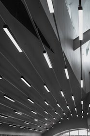 A series of modern, linear ceiling lights are suspended in an architectural interior. The ceiling features geometric shapes, and the lighting creates a pattern of elongated lines across the space. Soft natural light filters through the windows, adding to the minimalist and sleek design.