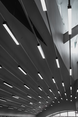 A series of modern, linear ceiling lights are suspended in an architectural interior. The ceiling features geometric shapes, and the lighting creates a pattern of elongated lines across the space. Soft natural light filters through the windows, adding to the minimalist and sleek design.
