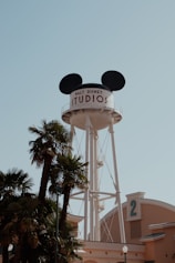 A tall water tower with large black mouse ears on top is labeled 'Walt Disney Studios.' It stands against a clear blue sky, surrounded by palm trees and next to a light brown building marked with the number 2.