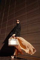 A person wearing a long flowing beige skirt and black outfit stands confidently against a backdrop of a modern, textured wall. This individual is accessorized with large sunglasses and is holding a stylish, structured handbag with a crocodile pattern. The dramatic play of light and shadow emphasizes the elegance of the attire and pose.