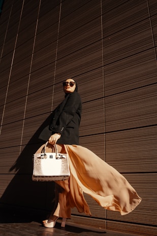 A person wearing a long flowing beige skirt and black outfit stands confidently against a backdrop of a modern, textured wall. This individual is accessorized with large sunglasses and is holding a stylish, structured handbag with a crocodile pattern. The dramatic play of light and shadow emphasizes the elegance of the attire and pose.