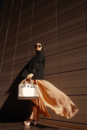A person wearing a long flowing beige skirt and black outfit stands confidently against a backdrop of a modern, textured wall. This individual is accessorized with large sunglasses and is holding a stylish, structured handbag with a crocodile pattern. The dramatic play of light and shadow emphasizes the elegance of the attire and pose.