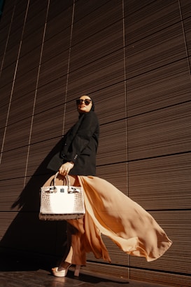 A person wearing a long flowing beige skirt and black outfit stands confidently against a backdrop of a modern, textured wall. This individual is accessorized with large sunglasses and is holding a stylish, structured handbag with a crocodile pattern. The dramatic play of light and shadow emphasizes the elegance of the attire and pose.