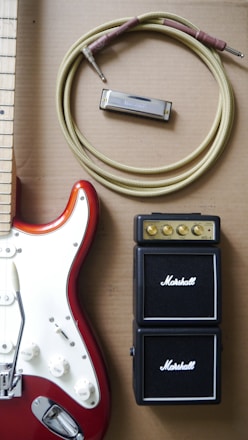 An electric guitar with a red and white body is positioned next to a coiled instrument cable, a harmonica, and a compact Marshall amplifier. The guitar features control knobs and metallic components, while the amplifier shows small golden dials for volume and tone control.