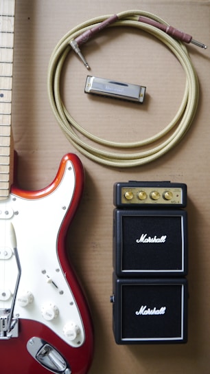 An electric guitar with a red and white body is positioned next to a coiled instrument cable, a harmonica, and a compact Marshall amplifier. The guitar features control knobs and metallic components, while the amplifier shows small golden dials for volume and tone control.