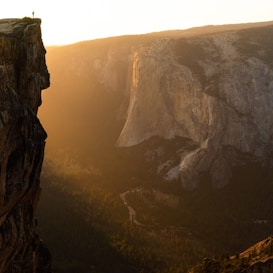 A breathtaking view of a vast canyon and rock formations, with the soft golden light of the setting sun casting long shadows and highlighting the rugged cliffs. A lone figure stands on the edge of a cliff, adding a sense of scale and human connection to the expansive natural landscape below.