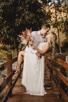 A couple shares an affectionate moment on a wooden bridge surrounded by lush greenery. The woman, dressed in a flowing white dress and holding a bouquet of flowers, is supported by the man, who wears a white shirt. They are close, almost kissing, creating a romantic and intimate atmosphere.