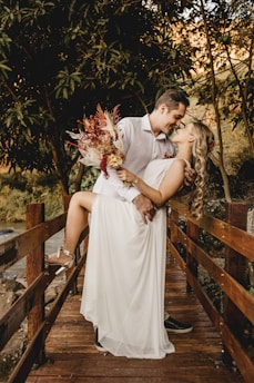 A couple shares an affectionate moment on a wooden bridge surrounded by lush greenery. The woman, dressed in a flowing white dress and holding a bouquet of flowers, is supported by the man, who wears a white shirt. They are close, almost kissing, creating a romantic and intimate atmosphere.