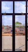 A construction site is viewed through a window with several panes. Workers are engaged in building activities, surrounded by piles of earth and construction materials. The sky is partly cloudy, and some greenery is visible in the background.