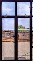 A construction site is viewed through a window with several panes. Workers are engaged in building activities, surrounded by piles of earth and construction materials. The sky is partly cloudy, and some greenery is visible in the background.
