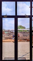 A construction site is viewed through a window with several panes. Workers are engaged in building activities, surrounded by piles of earth and construction materials. The sky is partly cloudy, and some greenery is visible in the background.