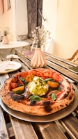 A wooden table in an outdoor setting holds a freshly prepared pizza with a thick, charred crust. The toppings include a large dollop of creamy burrata cheese, fresh basil leaves, yellow cherry tomatoes, and a drizzle of pesto sauce. In the background, there are tables with white plates and small vases holding dried flowers, adding a rustic and cozy ambiance.