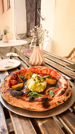 A wooden table in an outdoor setting holds a freshly prepared pizza with a thick, charred crust. The toppings include a large dollop of creamy burrata cheese, fresh basil leaves, yellow cherry tomatoes, and a drizzle of pesto sauce. In the background, there are tables with white plates and small vases holding dried flowers, adding a rustic and cozy ambiance.