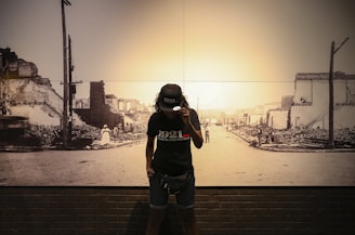 A person stands in front of a historic photograph depicting a street with damaged buildings and debris. The person wears a cap and a t-shirt with the year 1921 printed on it. The background photo shows an urban area that appears to have experienced destruction.