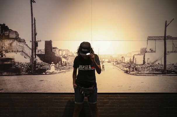 A person stands in front of a historic photograph depicting a street with damaged buildings and debris. The person wears a cap and a t-shirt with the year 1921 printed on it. The background photo shows an urban area that appears to have experienced destruction.