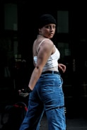 A person with a confident stance is wearing a black beanie, white sleeveless top, and blue high-waisted pants. The lighting highlights the person's features against a dark, urban background, creating a striking contrast.