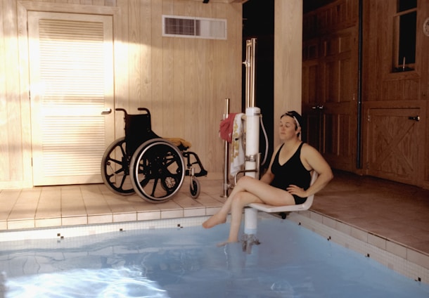 A woman in a black swimsuit is seated on a pool lift, preparing to enter a swimming pool. The setting is indoors with wooden paneling, and a wheelchair is positioned next to a door.