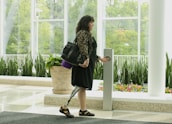 A woman with a prosthetic leg is standing indoors near a large window. She holds a handbag on her shoulder and is reaching out to press a button on a tall, sleek object in front of her. The room is decorated with green plants in planters and has a polished floor. Outside the window, there is lush greenery.