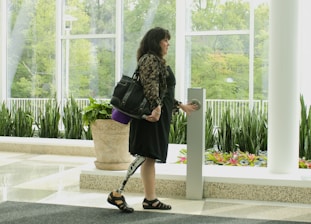 A woman with a prosthetic leg is standing indoors near a large window. She holds a handbag on her shoulder and is reaching out to press a button on a tall, sleek object in front of her. The room is decorated with green plants in planters and has a polished floor. Outside the window, there is lush greenery.