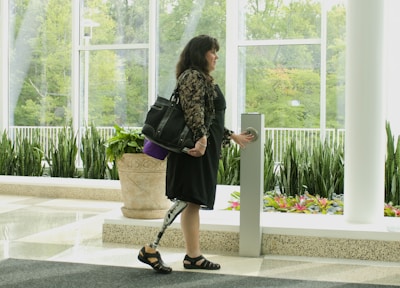 A woman with a prosthetic leg is standing indoors near a large window. She holds a handbag on her shoulder and is reaching out to press a button on a tall, sleek object in front of her. The room is decorated with green plants in planters and has a polished floor. Outside the window, there is lush greenery.