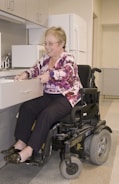 A person in a power wheelchair is smiling while opening a drawer in a kitchen. The kitchen features cabinets, a microwave, and a refrigerator. The person is wearing glasses and a pink patterned blouse with dark pants.