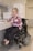 A person in a power wheelchair is smiling while opening a drawer in a kitchen. The kitchen features cabinets, a microwave, and a refrigerator. The person is wearing glasses and a pink patterned blouse with dark pants.