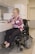 A person in a power wheelchair is smiling while opening a drawer in a kitchen. The kitchen features cabinets, a microwave, and a refrigerator. The person is wearing glasses and a pink patterned blouse with dark pants.