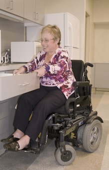 A person in a power wheelchair is smiling while opening a drawer in a kitchen. The kitchen features cabinets, a microwave, and a refrigerator. The person is wearing glasses and a pink patterned blouse with dark pants.