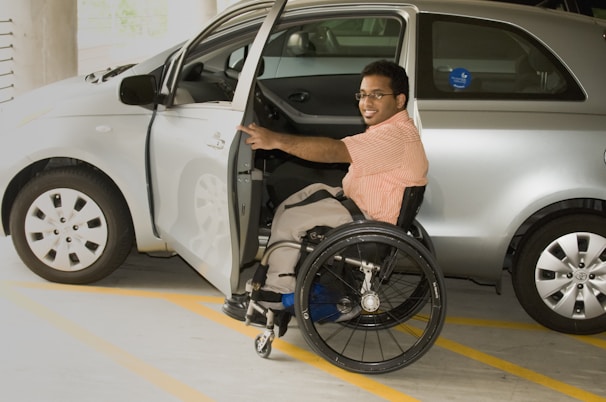 A person in a wheelchair is positioned next to an open car door, appearing to be preparing to transfer into or out of the vehicle. The car is parked in a designated parking space inside a parking garage. The individual is smiling and wearing glasses and a striped shirt.
