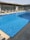 A clear swimming pool with sparkling blue water is surrounded by several empty sun loungers. The setting appears to be on a rooftop with a modern architectural design, featuring large glass panels and a pergola structure providing partial shade.