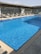 A clear swimming pool with sparkling blue water is surrounded by several empty sun loungers. The setting appears to be on a rooftop with a modern architectural design, featuring large glass panels and a pergola structure providing partial shade.