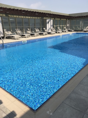 A clear swimming pool with sparkling blue water is surrounded by several empty sun loungers. The setting appears to be on a rooftop with a modern architectural design, featuring large glass panels and a pergola structure providing partial shade.