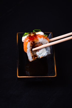 A sushi roll is held by chopsticks over a rectangular black dish. The sushi is topped with a slice of salmon, garnished with a dark sauce and slivers of green onion.