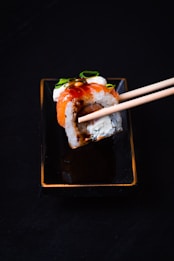 A sushi roll is held by chopsticks over a rectangular black dish. The sushi is topped with a slice of salmon, garnished with a dark sauce and slivers of green onion.