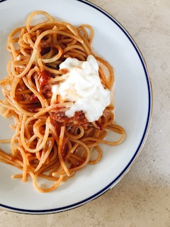 A plate of spaghetti topped with a tomato-based sauce and a dollop of creamy white cheese, set against a light-colored tabletop.