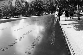 Engraved names are displayed in a solemn, memorial setting with trees and a reflective surface. People are walking nearby, and the area appears to be outdoors, suggesting a commemorative site.