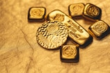 A collection of gold bullion including bars and coins, featuring intricate designs and inscriptions visible on the surfaces. The items are placed on a textured golden background, emphasizing their shiny and luxurious appearance.