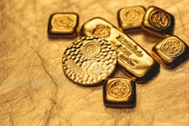 A collection of gold bullion including bars and coins, featuring intricate designs and inscriptions visible on the surfaces. The items are placed on a textured golden background, emphasizing their shiny and luxurious appearance.