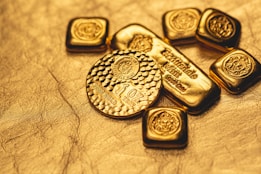 A collection of gold bullion including bars and coins, featuring intricate designs and inscriptions visible on the surfaces. The items are placed on a textured golden background, emphasizing their shiny and luxurious appearance.