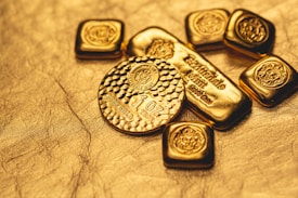 A collection of gold bullion including bars and coins, featuring intricate designs and inscriptions visible on the surfaces. The items are placed on a textured golden background, emphasizing their shiny and luxurious appearance.